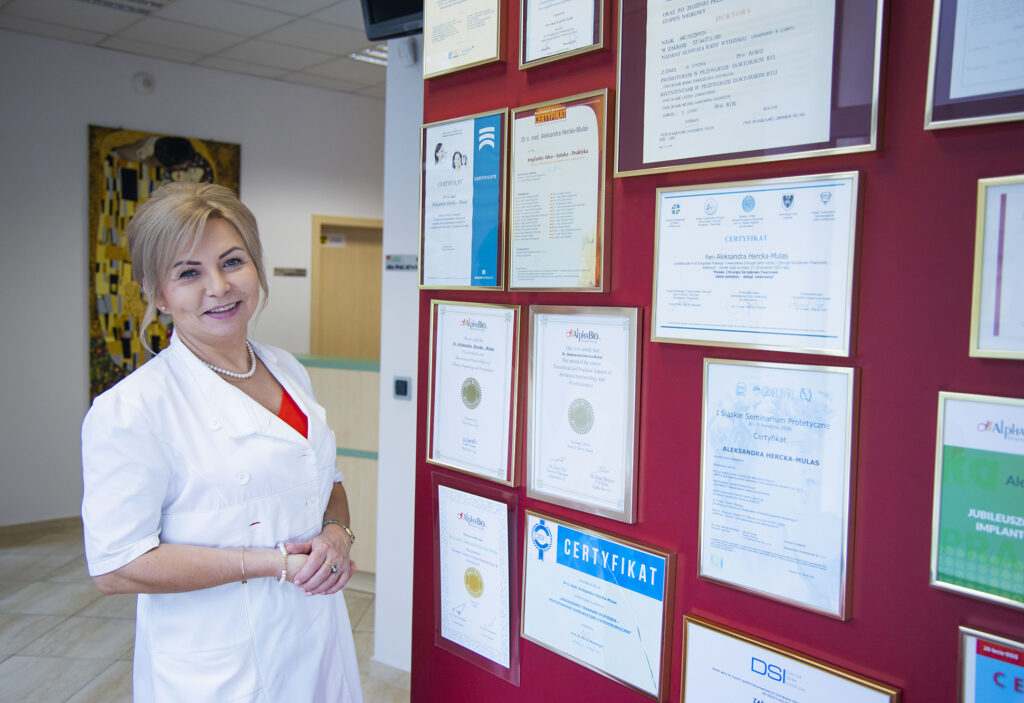 Dr Hercka przy ścianie certyfikatów i dyplomów